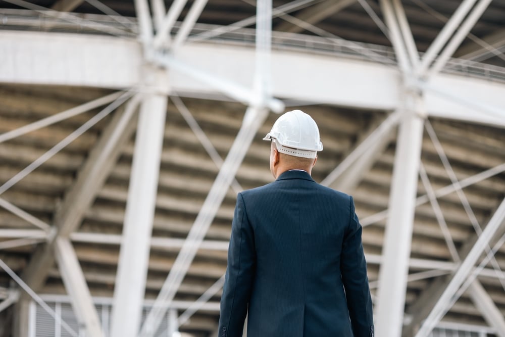 Back view of a professional in a suit and white hard hat surveying a steel construction framework.