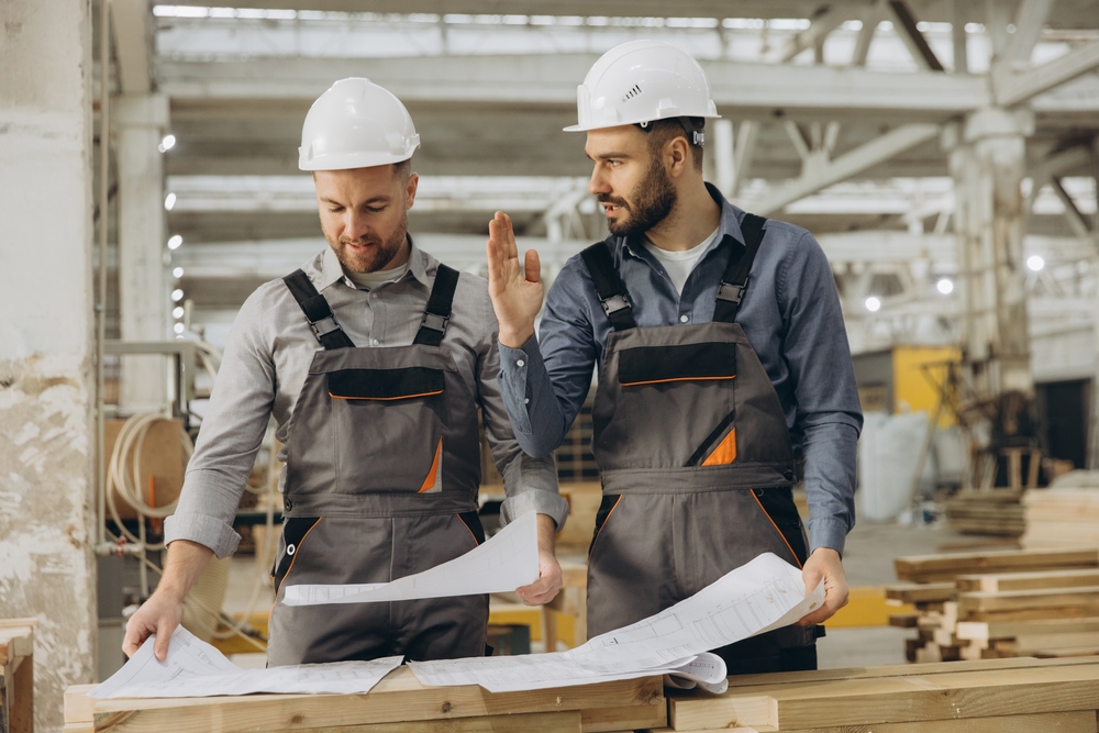 Twobuilderswearinghardhatsreviewingblueprintsinamodularbuilding | engco Two Construction Workers in Hard Hats Examine Blueprints on a Wooden Workbench