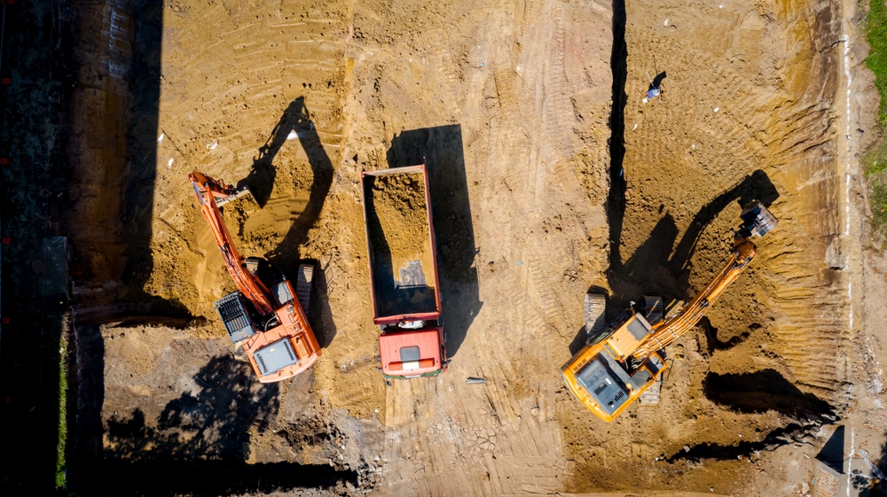 Abovetopviewontwobigexcavatorsastheydigging | engco a Large Cleared Construction Site with Dirt Piles and Heavy Equipment Next to a Suburban Neighborhood to the Right and Trees to the Left