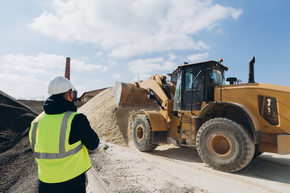 Construction worker in a neon vest and hard hat watches a large loader dump material onto a pile on a gravel site.