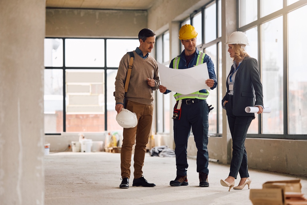 Constructionworkerexaminingblueprintswitharchitectsatbuildingsitecopy | engco Three Construction Professionals Review Blueprints Inside a Partly Finished Building Inside a Bright Space