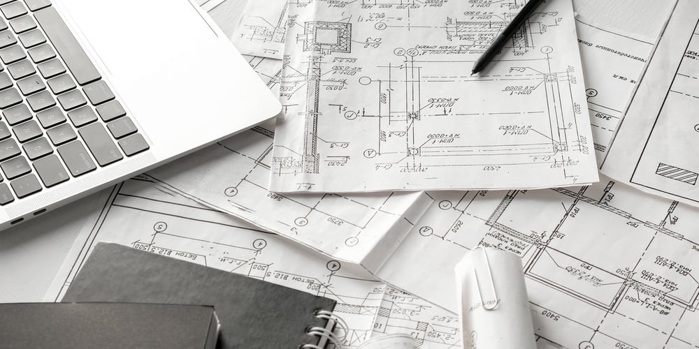 Architectural blueprints spread over a desk with a laptop, pencil, and notebook for drafting.